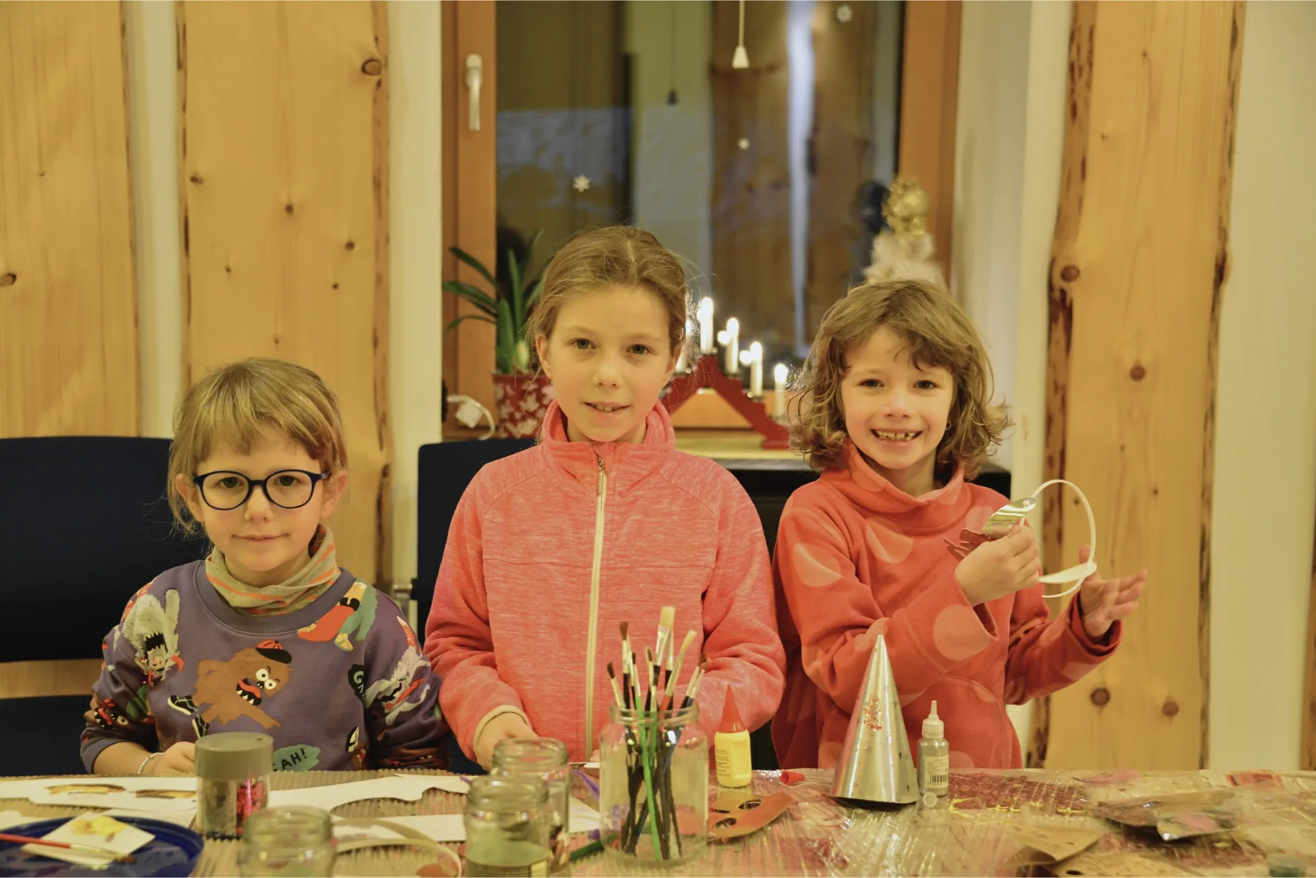 Tři děti při vánočním tvoření v Montanie Resort v Jizerských horách, u stolu s výtvarnými potřebami v útulném dřevěném interiéru.
