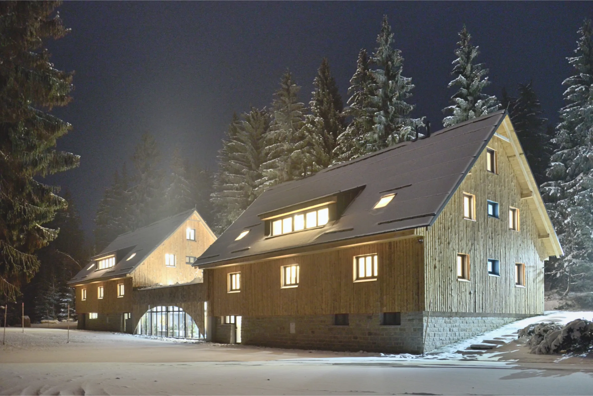 Noční pohled na osvětlené dřevěné budovy resortu Montanie zasazené do zasněženého smrkového lesa v Jizerských horách.