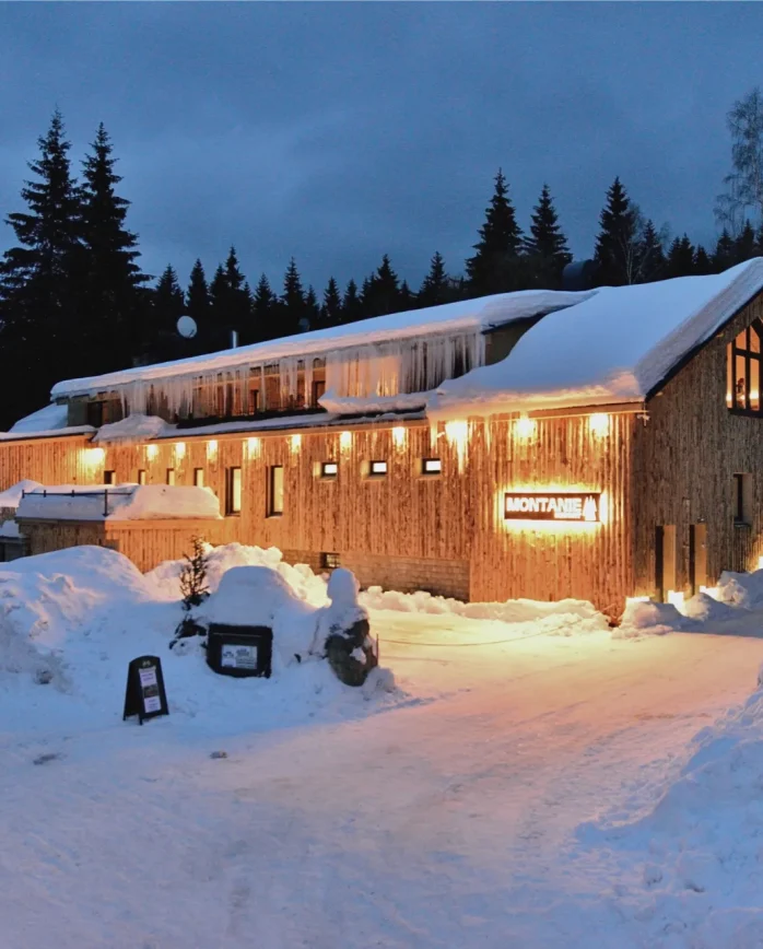 Zasněžený Montanie Resort v Jizerských horách za zimního soumraku, s osvětlenou dřevěnou budovou a běžkaři na sněhové cestě.
